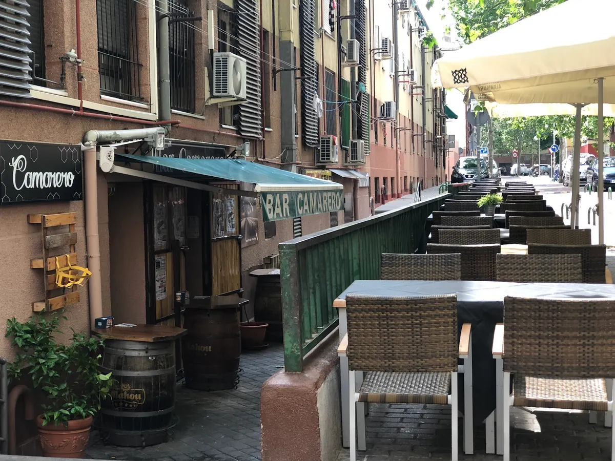 Entrada del bar con el toldo verde «Bar Camarero», barricas en la puerta y la terraza a lo largo del Pasaje de la Rueca
